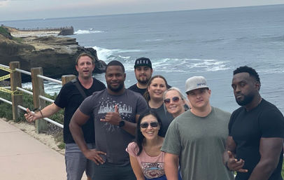 Eight people posing on an oceanfront trail along rocky coastal cliffs with a railing, crashing waves and an overcast sky
