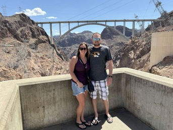 Smiling couple in casual summer clothes pose on a concrete overlook at Hoover Dam, with the arched memorial bridge and rocky desert canyon under a clear blue sky.