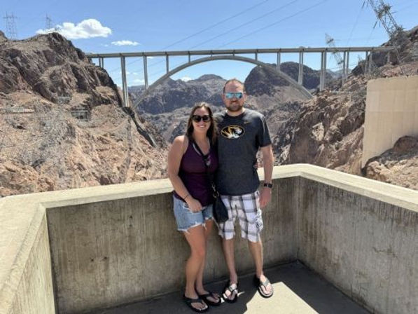 Smiling couple in casual summer clothes pose on a concrete overlook at Hoover Dam, with the arched memorial bridge and rocky desert canyon under a clear blue sky.
