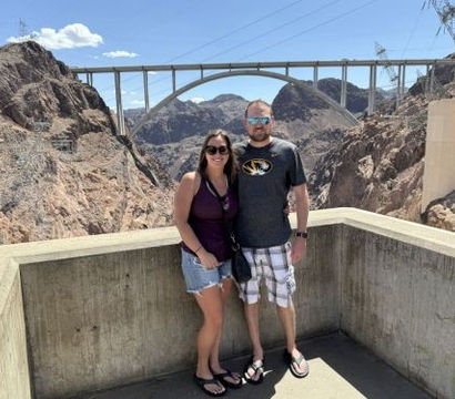 Smiling couple in casual summer clothes pose on a concrete overlook at Hoover Dam, with the arched memorial bridge and rocky desert canyon under a clear blue sky.