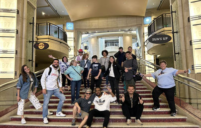 Playful group photo of about 15 people posing and waving on a wide carpeted grand staircase inside a bright hotel or theater lobby with balconies and metal railings.