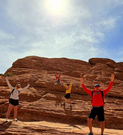 Four hikers with backpacks cheering on layered red sandstone rocks beneath a bright sun and blue sky — sunny red-rock desert outdoor adventure.