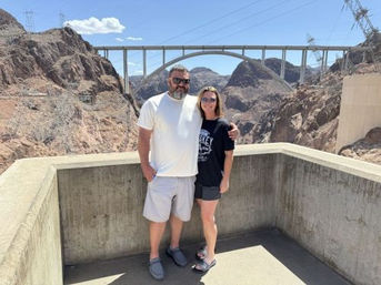 Two people posing on a concrete overlook at Hoover Dam with the arched bridge and rocky desert canyon under a bright blue sky