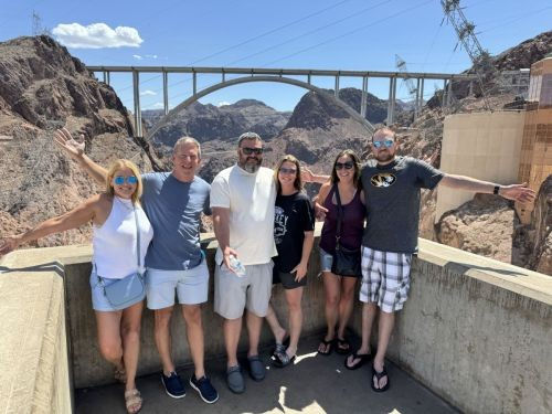 Six adults posing on a Hoover Dam overlook with arms outstretched, arch bridge and rocky Nevada–Arizona canyon in the background under a bright blue sky.