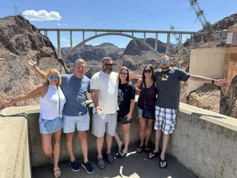 Six adults posing on a Hoover Dam overlook with arms outstretched, arch bridge and rocky Nevada–Arizona canyon in the background under a bright blue sky.