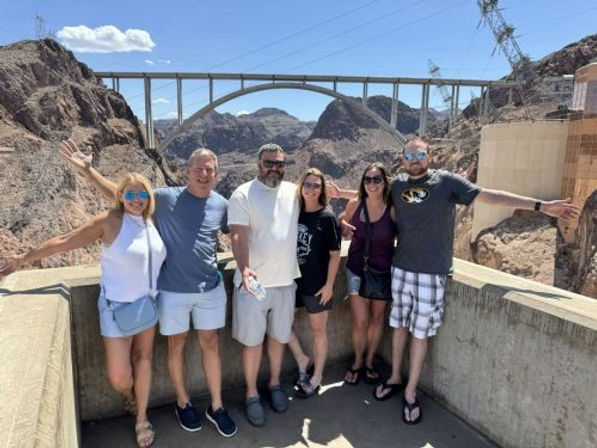 Six adults posing on a Hoover Dam overlook with arms outstretched, arch bridge and rocky Nevada–Arizona canyon in the background under a bright blue sky.