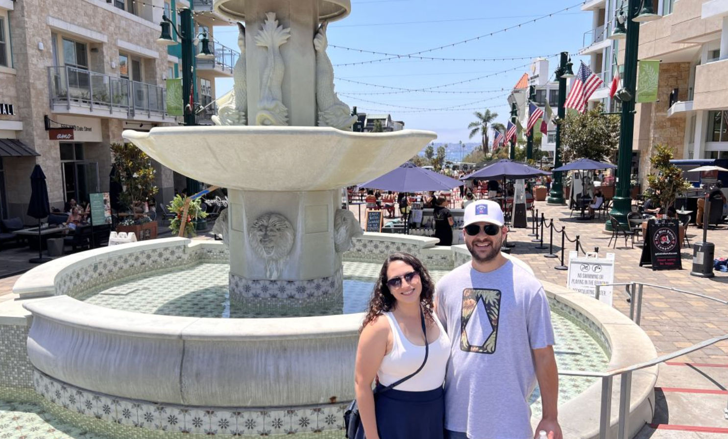 Couple posing by an ornate stone fountain on a sunny beachfront promenade with outdoor dining umbrellas, American flags, palm trees and distant ocean views.