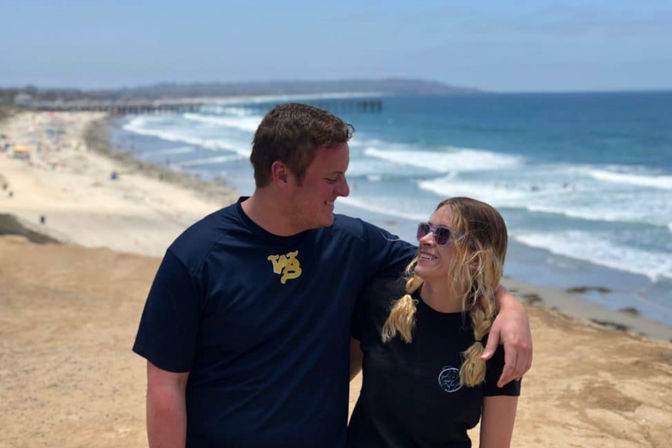 Smiling couple on a coastal bluff with an arm around each other, overlooking a sandy beach, rolling ocean waves and a distant pier on a sunny day.