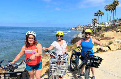 Three helmeted cyclists on electric bikes posing on a sunny oceanfront cliffside path with palm trees, coastal houses and the blue Pacific Ocean in the background.