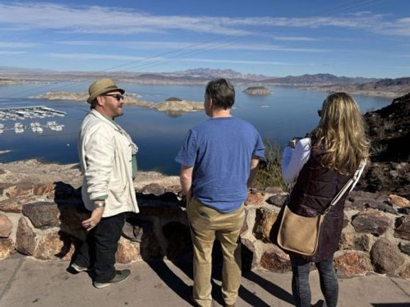 Three visitors at a rocky overlook gaze over a marina and wide blue desert lake with small islands and distant mountains under a clear sky — panoramic scenic lake overlook.