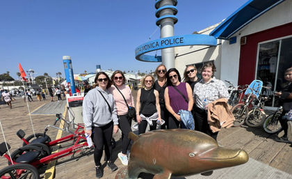 Smiling group of people posing on a sunny wooden oceanfront pier beside a bronze dolphin sculpture, with rental bikes, tricycles and a blue police kiosk visible in the background.