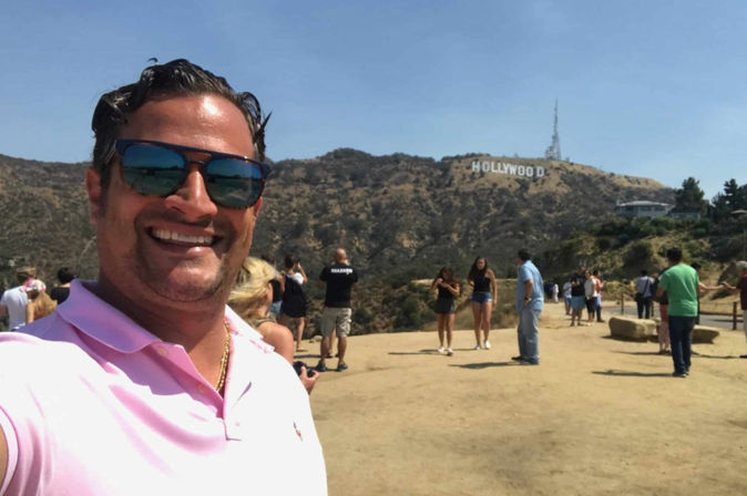 Person in sunglasses taking a selfie at the Hollywood Sign overlook in Los Angeles, with tourists on a dusty trail and rolling Hollywood Hills under a clear blue sky.