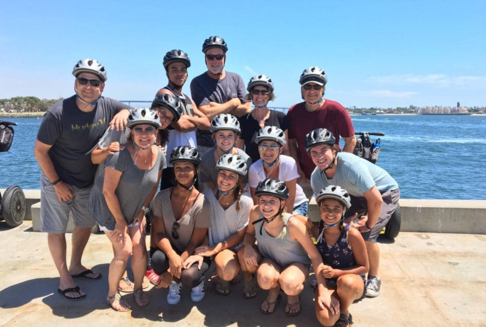Smiling group of adults and teens wearing helmets posing on a sunny coastal pier by the blue ocean with Segways visible behind them