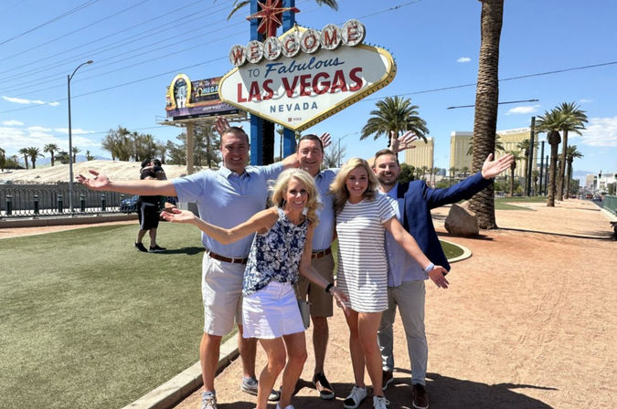 Five adults smiling and posing with arms outstretched at the iconic "Welcome to Fabulous Las Vegas, Nevada" sign on a sunny day with palm trees and Strip hotels in the background.
