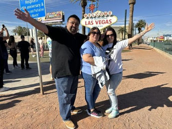 Group of three smiling people posing with arms outstretched in front of the iconic Welcome to Fabulous Las Vegas sign in Las Vegas, Nevada, with palm trees and tourists in the sunny background.