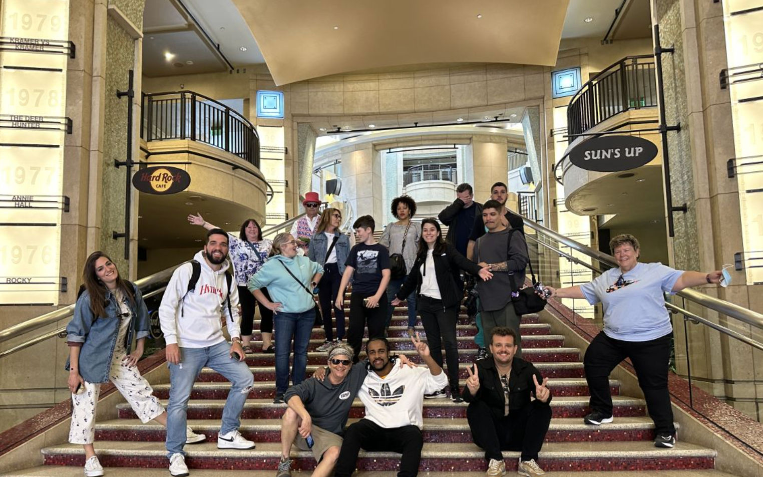 Cheerful group of about 15 people casually posing on a wide red-carpeted staircase inside a grand theater-style lobby with balconies, stone pillars displaying year plaques and decorative signage.