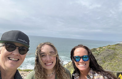 Three smiling people taking a coastal selfie at a cliffside overlook with blue-gray ocean waves, cloudy sky, and scrubby shoreline in the background.