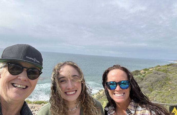 Three smiling people taking a coastal selfie at a cliffside overlook with blue-gray ocean waves, cloudy sky, and scrubby shoreline in the background.