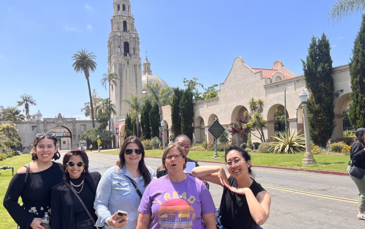 Cheerful group of six adults posing in front of Balboa Park’s California Tower and arched Spanish-style buildings in San Diego under a clear blue sky.