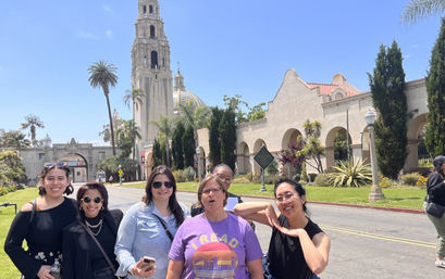 Cheerful group of six adults posing in front of Balboa Park’s California Tower and arched Spanish-style buildings in San Diego under a clear blue sky.