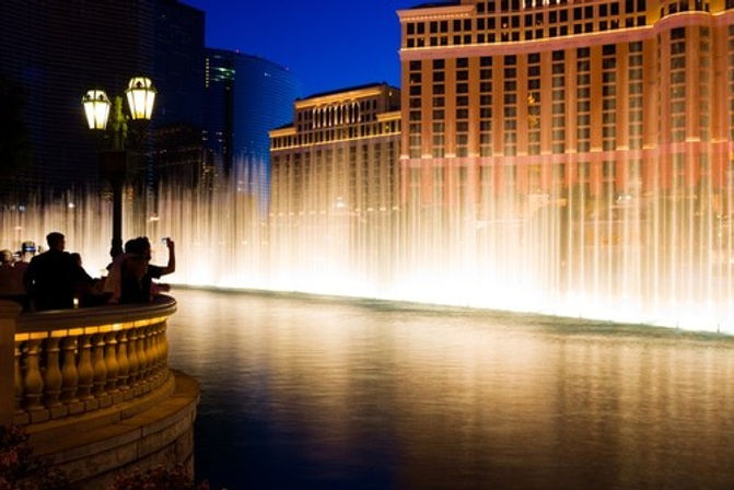 Nighttime hotel fountain show with illuminated water jets rising across a reflective pool, warm-lit hotel facade in the background and silhouetted visitors photographing the spectacle from a curved balustrade.