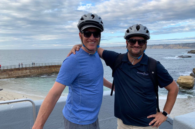 Two friends wearing bicycle helmets and sunglasses smiling with arms around each other on an oceanfront promenade overlooking rocky cliffs, surf, and a cloudy sky.