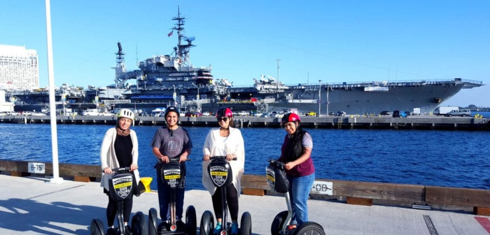 Four people on Segways wearing helmets on a sunny waterfront pier with a docked naval aircraft carrier and harbor in the background.