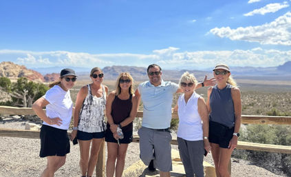 Smiling group of six adults in casual hiking clothes and sunglasses posing at a wooden railing on a sunny desert canyon overlook with red-rock formations and a distant mountain vista under a bright blue sky