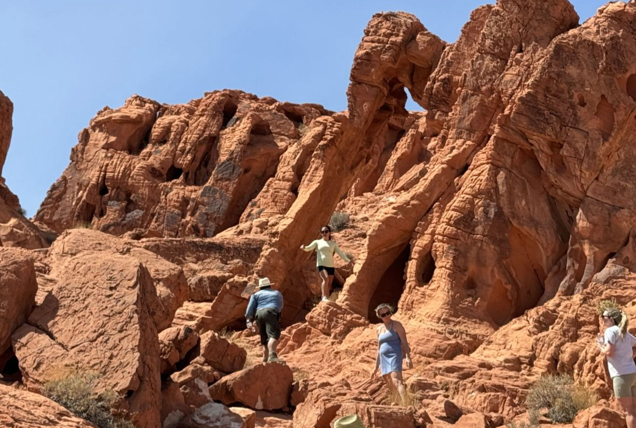 Group of hikers scrambling up sunlit red sandstone cliffs with a small natural arch and a clear blue desert sky