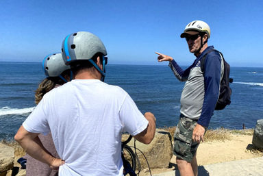 Three cyclists in helmets on a sunny coastal bluff — a guide points toward the ocean waves on a scenic seaside bike tour