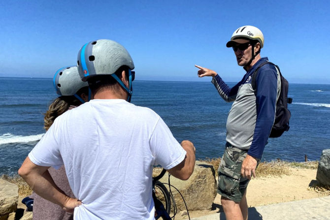 Three cyclists in helmets on a sunny coastal bluff — a guide points toward the ocean waves on a scenic seaside bike tour