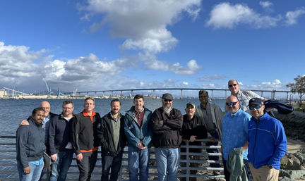 Group of men posing at a waterfront railing with a long arched harbor bridge and city skyline under a bright blue sky with puffy clouds.