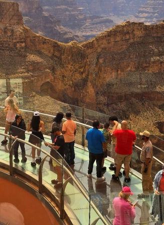 Visitors on a curved glass skywalk above a vast orange-brown desert canyon with dramatic layered cliffs — Arizona scenic overlook
