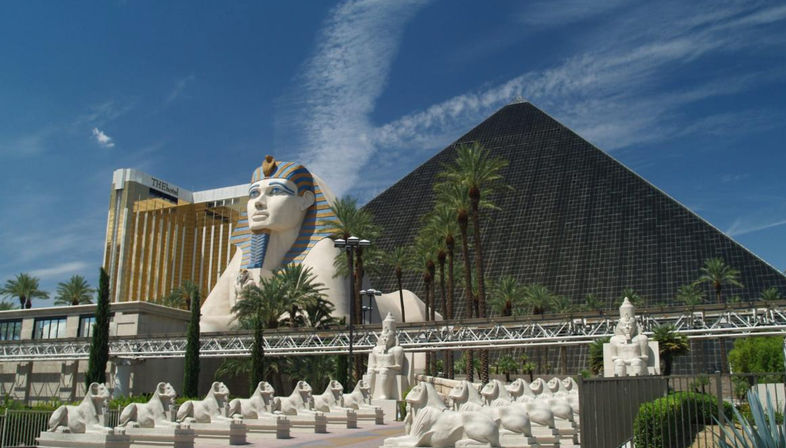 Black glass pyramid hotel with a giant Egyptian sphinx replica, rows of small sphinx statues and palm trees under a bright blue sky on the Las Vegas Strip.