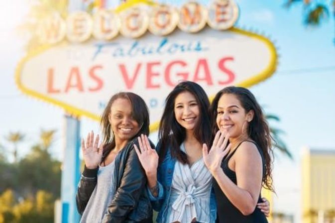 Three smiling women waving in front of the iconic 'Welcome to Fabulous Las Vegas' sign on a sunny day