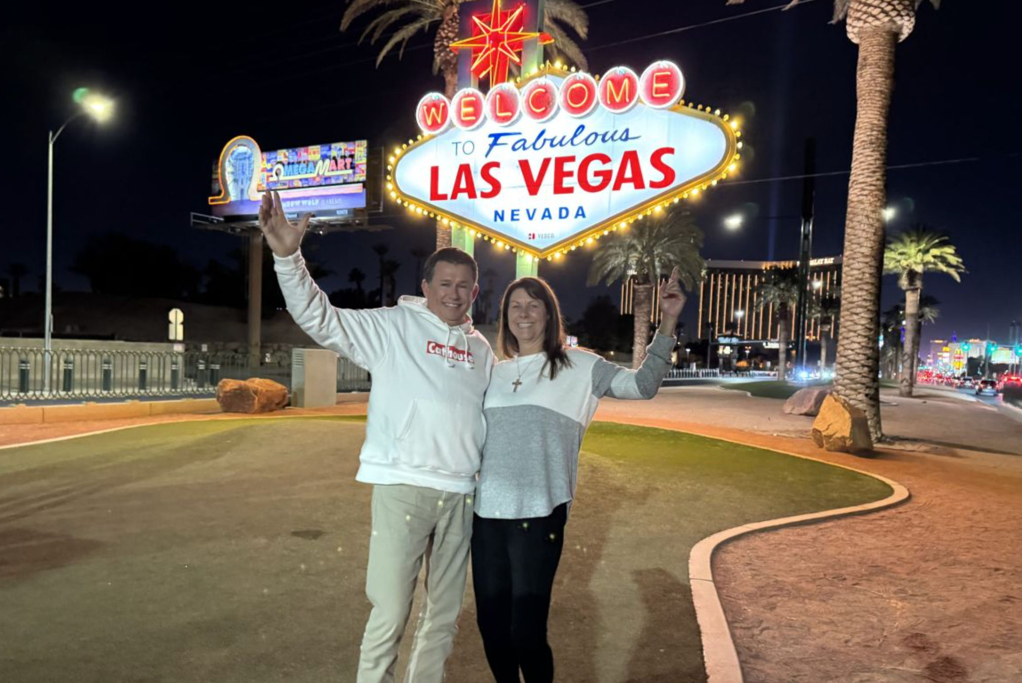 Two smiling tourists pose at night beneath the illuminated Welcome to Fabulous Las Vegas Nevada sign, with palm trees and bright Strip lights in the background.