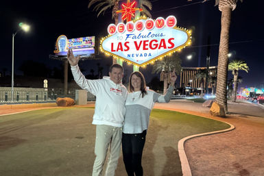 Two smiling tourists pose at night beneath the illuminated Welcome to Fabulous Las Vegas Nevada sign, with palm trees and bright Strip lights in the background.