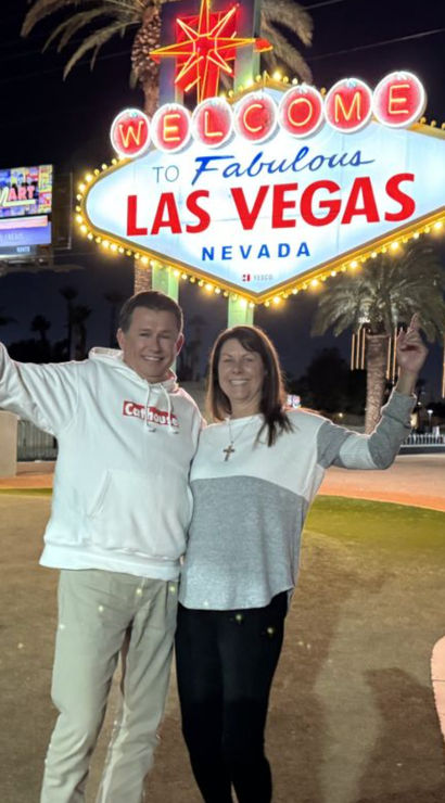 Two smiling tourists pose at night beneath the illuminated Welcome to Fabulous Las Vegas Nevada sign, with palm trees and bright Strip lights in the background.