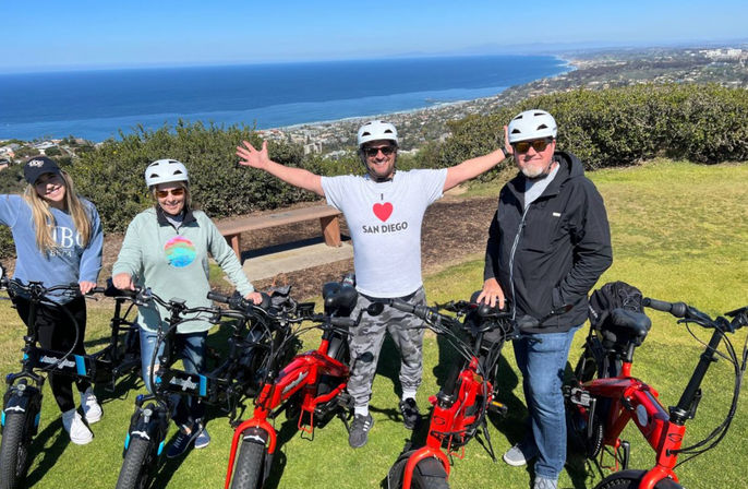 Four people in helmets posing with red and black electric bikes on a grassy hilltop lookout, central person wearing an "I ♥ San Diego" shirt with arms outstretched, panoramic Pacific Ocean coastline and city below.