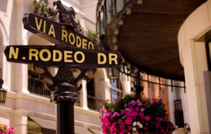 Black-and-gold street signs reading "Via Rodeo" and "N. Rodeo Dr" on an ornamental post in a chic upscale shopping district, with pink hanging flowers and elegant building facades