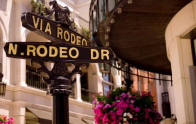 Black-and-gold street signs reading "Via Rodeo" and "N. Rodeo Dr" on an ornamental post in a chic upscale shopping district, with pink hanging flowers and elegant building facades