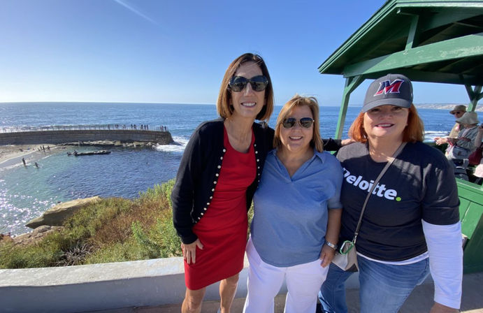Three smiling women in sunglasses posing at a sunny coastal viewpoint overlooking a seawall and tide pools with the blue ocean and clear sky behind them and a green wooden shelter at right.