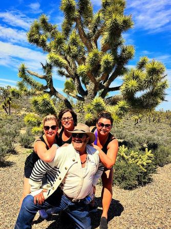 Four friends wearing sunglasses pose playfully in front of a large Joshua tree under a bright blue sky in the Mojave Desert desert landscape.