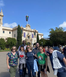 Group of seven adults smiling and posing in front of a historic Spanish‑style museum facade with tall cypress trees and a colorful flag under a bright blue sky in Balboa Park, San Diego.