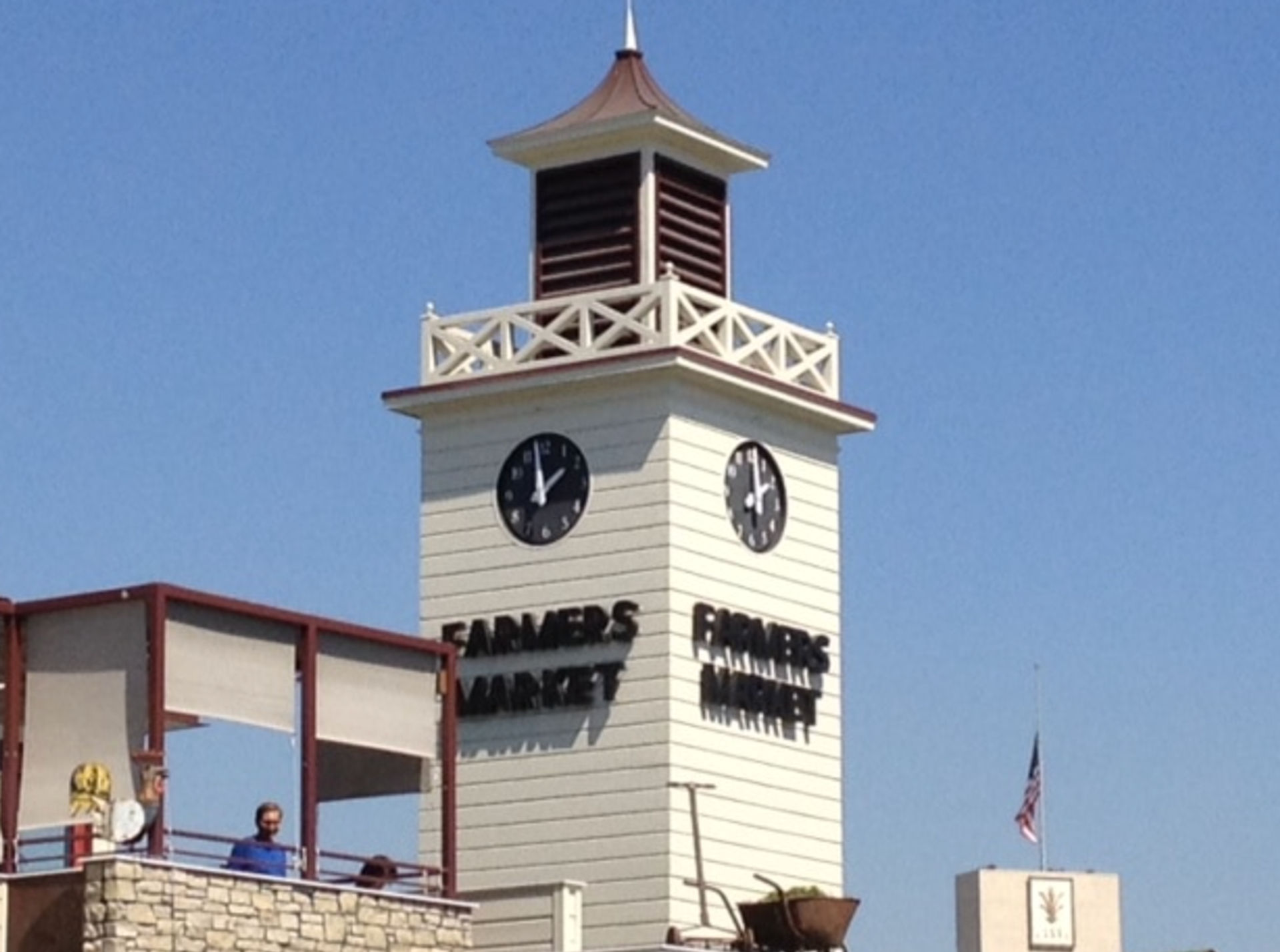 White clock tower labeled "Farmers Market" with two black clock faces above a sunny outdoor market terrace and American flag