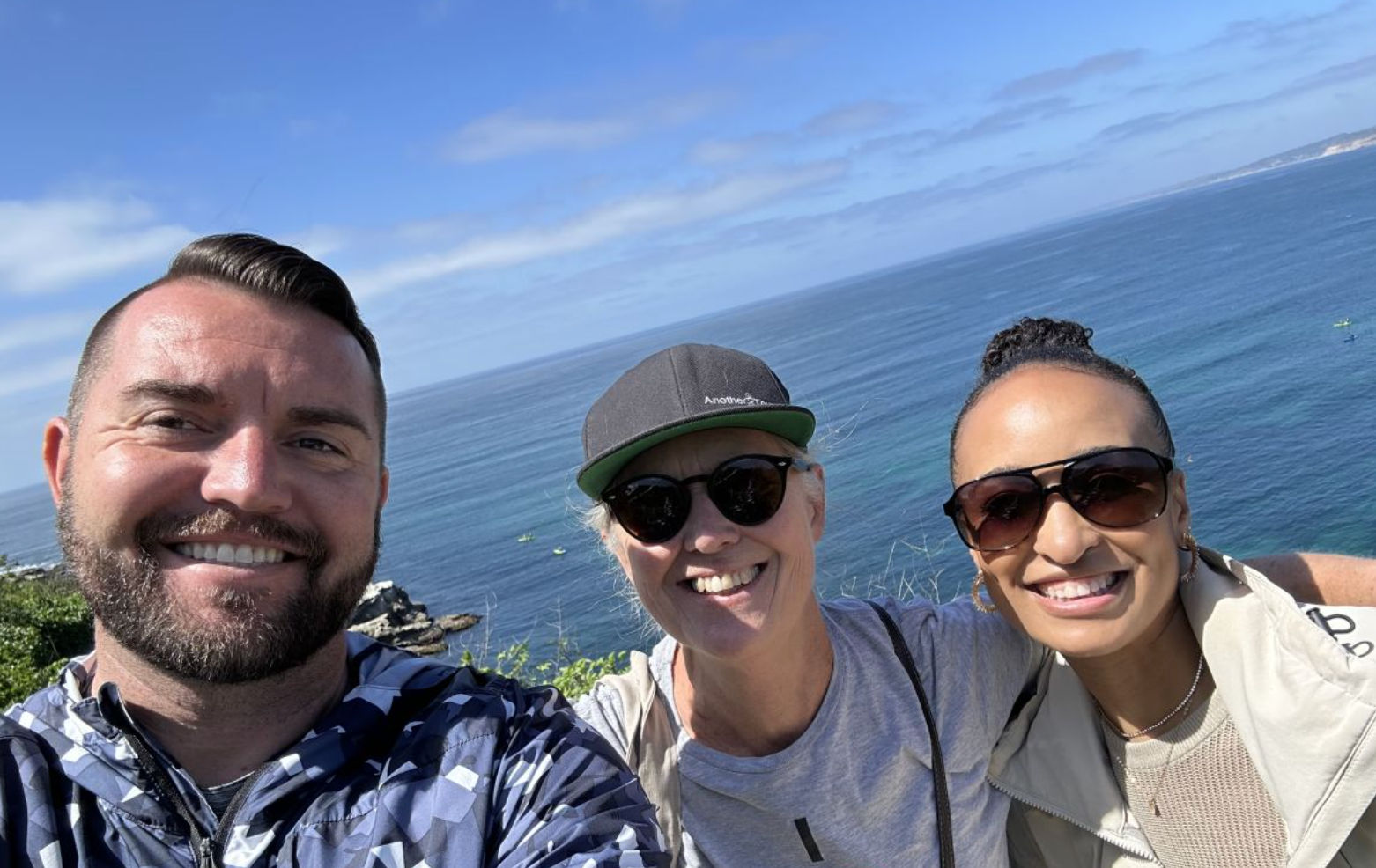 Three smiling people taking a sunny seaside selfie on a coastal bluff, wearing sunglasses and a cap with deep blue ocean and distant coastline in the background.