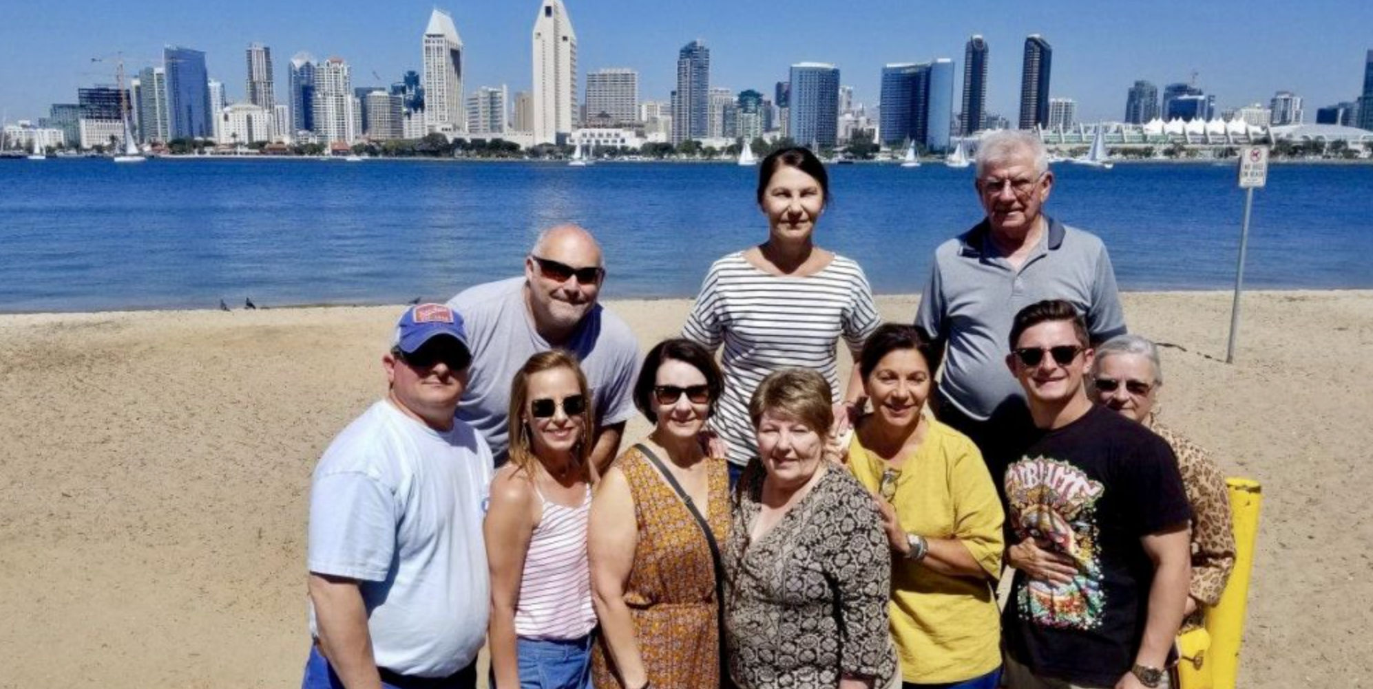 Multigenerational group posing on a sunny sandy beach with the San Diego skyline and bay in the background and sailboats on the water.