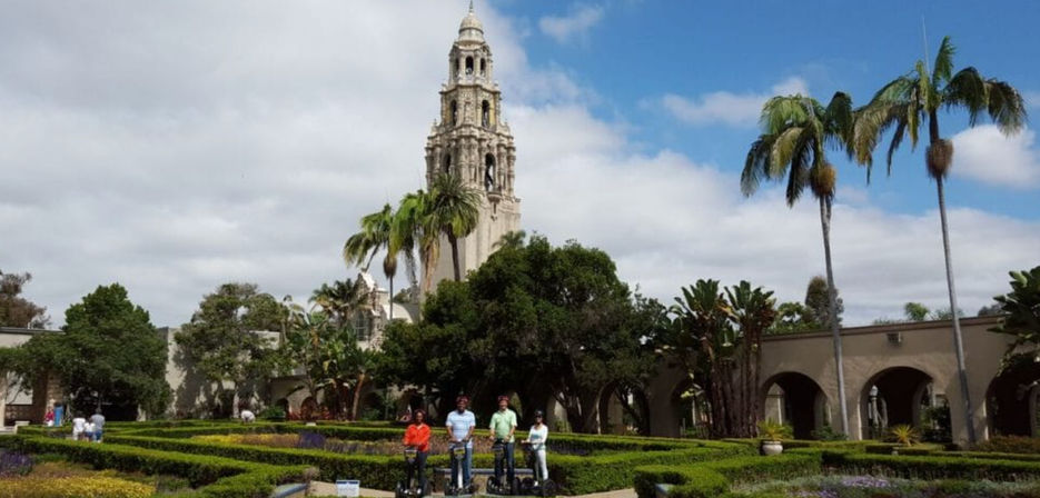 Sunny formal garden with hedged pathways and palm trees, a historic Spanish-style bell tower in the background, and four people riding Segways in the foreground.