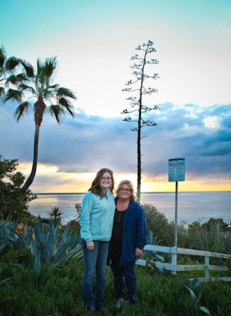 Two smiling women posing on a coastal bluff at sunset with palm trees, agave plants, and a calm ocean under a colorful sky