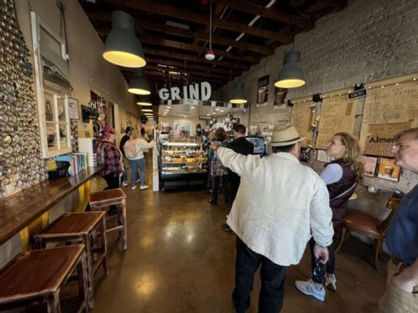 Cozy rustic coffee shop interior with pastry display case, hanging pendant lights, wooden beams, bar seating and patrons queuing.